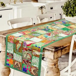 May include: A rectangular table runner featuring a patchwork design of various green, yellow, and multi-colored fabric squares with intricate floral and paisley embroidery. The runner has a solid green border and the text @jaipurarts printed on it.