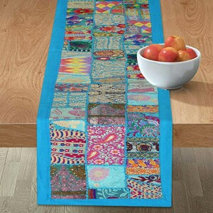 Può includere: Un runner da tavola rettangolare caratterizzato da un vivace patchwork di vari quadrati di tessuto colorati con motivi complessi, bordato da un tessuto blu brillante a tinta unita, accompagnato da una ciotola bianca con mele rosse e gialle.