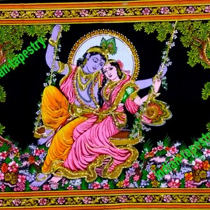 K&ouml;nnte beinhalten: Eine farbenfrohe Wandteppich, der ein Paar auf einer Schaukel in einem &uuml;ppigen gr&uuml;nen Wald zeigt. Das Paar ist in traditioneller indischer Kleidung gekleidet, die Frau tr&auml;gt ein rosa und gr&uuml;nes Sari und der Mann einen blauen und gelben Dhoti. Der Wandteppich ist mit einem goldenen Rahmen versehen und weist komplizierte Details auf.