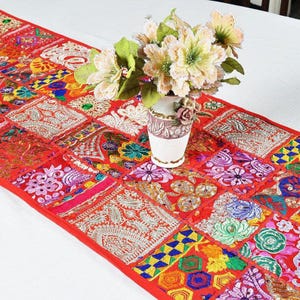 May include: A long rectangular table runner featuring a patchwork design of red fabric squares adorned with multi-coloured embroidery, sequins, and metallic thread patterns. A white ceramic vase containing a bouquet of light-coloured flowers sits on top.