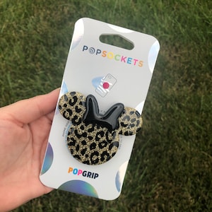 May include: A black and gold PopSockets PopGrip featuring a Minnie Mouse design with a leopard print pattern. The PopGrip is attached to a white and silver PopSockets branded backing card. The PopGrip is designed to attach to a mobile phone.