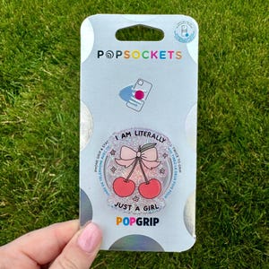 May include: A PopSockets PopGrip with a cherry design and the text "I AM LITERALLY JUST A GIRL". The grip is pink and glittery, with a bow and two cherries. The PopSockets packaging is white and silver.