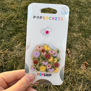 May include: A PopSockets PopGrip phone grip with a floral design. The grip features a variety of flowers in shades of pink, purple, red, and yellow, with a gold glitter background. The text "PopGrip" is printed in white on the grip.