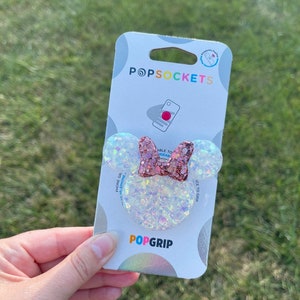 May include: A PopSockets PopGrip featuring a Minnie Mouse design. The grip has a clear, glittery base with two round ears and a pink glitter bow. The PopSockets packaging is white with the brand name and a phone graphic.