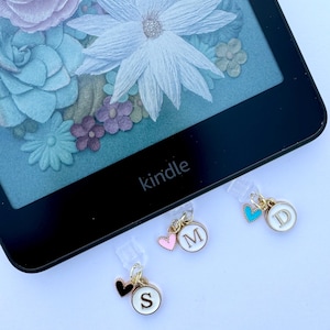 Może przedstawiać: Trzy złote breloki do zakładek z czarnymi, różowymi i niebieskimi sercami oraz literami "S", "M" i "D" na białych kółkach. Breloki są przymocowane do czarnego czytnika Kindle.