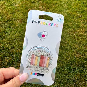 May include: A PopSockets PopGrip with a clear design featuring a stack of books in pastel colors and floral accents. The text reads "THERE'S NO SUCH THING as too many books." The PopSockets packaging is white with the brand name and logo.