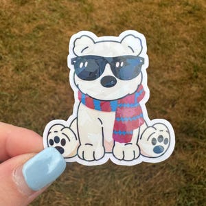 May include: A white polar bear sticker wearing sunglasses and a red, blue, and white scarf. The bear is sitting with its paws forward. The sticker is on a white background with a brown background.