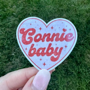 Może przedstawiać: Naklejka w kształcie serca z napisem "Connie baby" w czerwonym, stylizowanym kroju pisma. Naklejka ma białą obw&oacute;dkę i jasnor&oacute;żowe tło z małymi akcentami gwiazd. Naklejka jest trzymana na tle zielonej trawy.