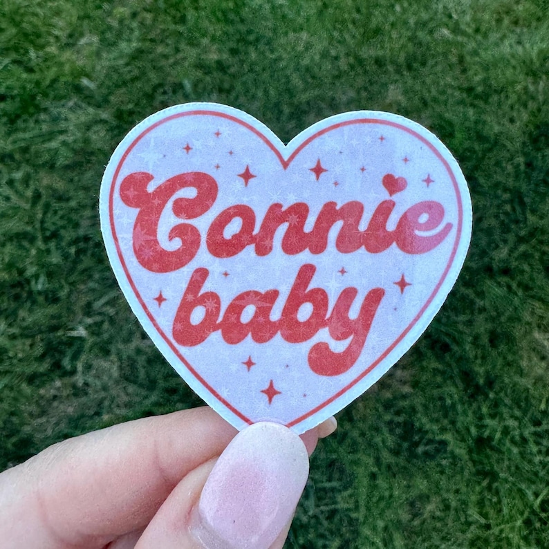 Może przedstawiać: Naklejka w kształcie serca z napisem "Connie baby" w czerwonym, stylizowanym kroju pisma. Naklejka ma białą obw&oacute;dkę i jasnor&oacute;żowe tło z małymi akcentami gwiazd. Naklejka jest trzymana na tle zielonej trawy.