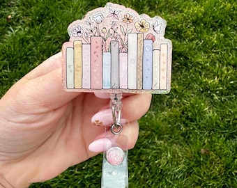 May include: A badge reel featuring a colorful illustration of a stack of books with flowers and butterflies on top. The badge reel has a clear plastic holder and a retractable cord. The design is glittery and has pastel colors.