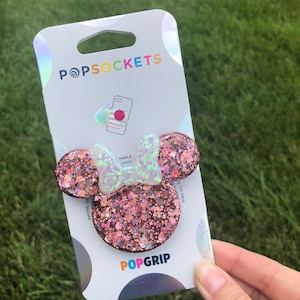 May include: A pink and white glitter PopSocket phone grip with a Minnie Mouse design. The PopSocket is on a white card with the words "PopSockets" and "PopGrip" printed on it.