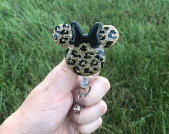 May include: A black and gold glitter leopard print Minnie Mouse head with a black bow, attached to a clear retractable badge reel.