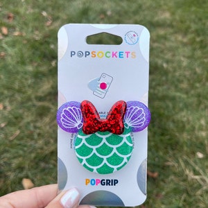 May include: A green, sparkly mermaid-themed PopGrip phone grip with purple and red glitter accents. The grip features a red bow and purple seashells. The packaging says "PopSockets" and "PopGrip".