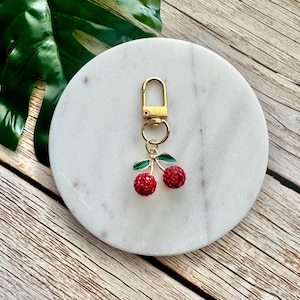May include: A gold-toned keychain featuring a pair of red, crystal-studded cherries with green leaves. The keychain is attached to a gold clasp. The cherries are the focal point, set against a white marble coaster and a wooden surface.