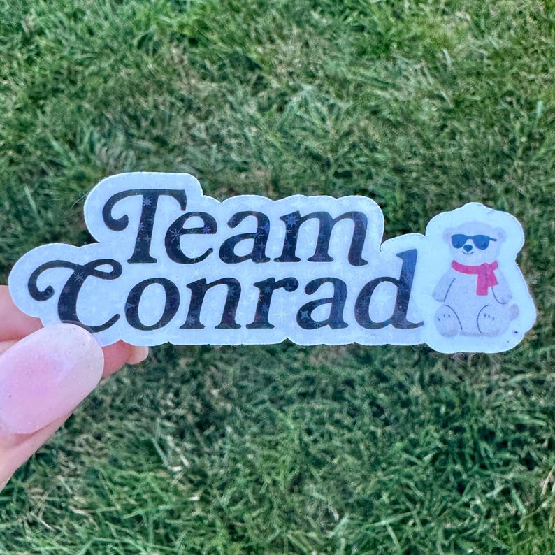 Pode incluir: Um adesivo com o texto "Team Conrad" em escrita preta. Um urso de desenho animado usando &oacute;culos de sol e um cachecol rosa est&aacute; no lado direito. O adesivo &eacute; segurado por uma m&atilde;o contra um fundo de grama verde.