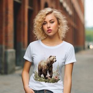 May include: White t-shirt with a brown bear walking through a field of flowers. The bear is surrounded by green foliage and pink flowers.