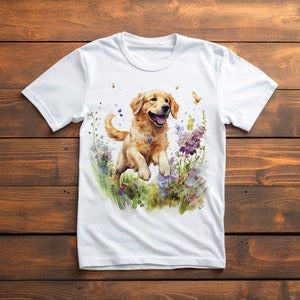 May include: White t-shirt featuring a watercolor illustration of a Golden Retriever dog jumping through a field of flowers. The dog is light brown with a happy expression. Butterflies and colorful flowers are also depicted.