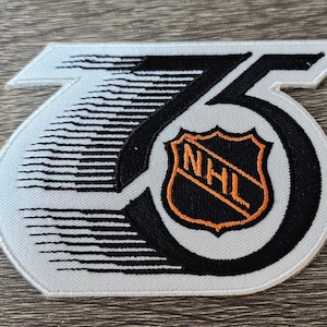 May include: A white and black embroidered patch featuring the number 75 and the NHL logo in orange and black.