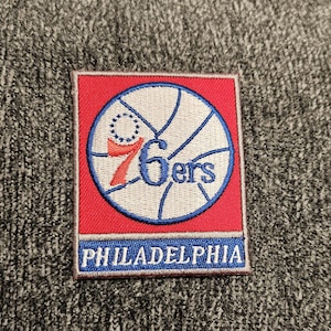 Könnte beinhalten: Ein gestickter Aufnäher mit einem Basketball und dem Text "76ers" und "PHILADELPHIA" in Rot und Blau.