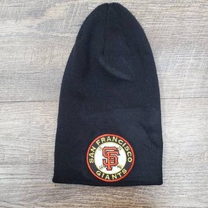 May include: Black beanie with a San Francisco Giants logo embroidered on the front. The logo is a circular patch with the team's name and a baseball with the letter "S" inside.