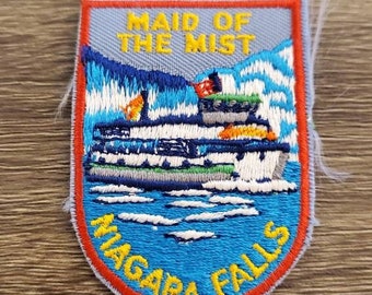 May include: Embroidered patch depicting a boat on water, with the text "MAID OF THE MIST" and "NIAGARA FALLS". The patch has a blue background, a red border, and yellow text. The design includes mountains and water.