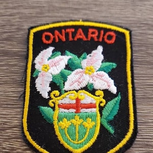 Puede incluir: Un parche bordado negro y amarillo con la palabra "ONTARIO" en letras rojas. El parche presenta un escudo verde y amarillo con una cruz blanca y tres flores blancas con centros rosados.