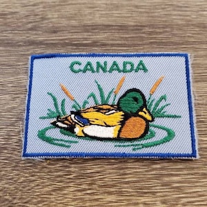 Puede incluir: Un parche bordado azul y blanco con la palabra "CANADA" en letras verdes. El parche presenta un pato amarillo con cabeza verde nadando en un estanque con juncos verdes.