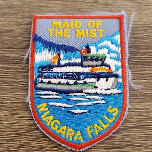 Puede incluir: Parche bordado con un barco en el agua, con el texto "MAID OF THE MIST" y "NIAGARA FALLS". El parche tiene un fondo azul, un borde rojo y texto amarillo. El diseño incluye montañas y agua.