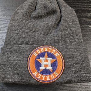 May include: A dark grey beanie hat with a circular patch featuring the Houston Astros logo. The patch has an orange border, a blue star, and the team's name in white lettering.