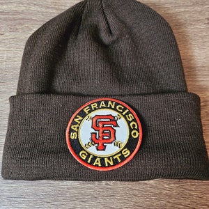 Może przedstawiać: Czarna czapka z dzianiny z okrągłą naszywką przedstawiającą logo San Francisco Giants w kolorze pomarańczowym, białym i czarnym. Naszywka zawiera napis "San Francisco Giants" wokół logo.