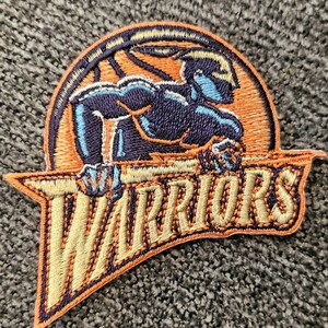 Op de afbeelding: Een geborduurde patch met het logo van de Golden State Warriors. Het logo is een gestileerd silhouet van een krijger die een basketbal vasthoudt, met het woord "WARRIORS" eronder geschreven in een gebogen schrift.
