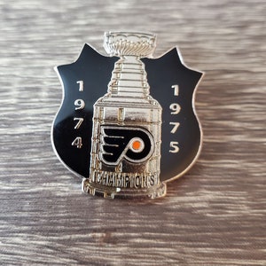 Op de afbeelding: Een zilveren en zwarte emaille pin met het logo van de Philadelphia Flyers en de Stanley Cup. De pin heeft de tekst "1974 1975 CHAMPIONS".