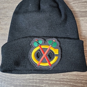 May include: Black knit beanie featuring a patch with a logo of a yellow 'C' with crossed red tomahawks, outlined in black, and green and black details. The beanie is folded at the brim.