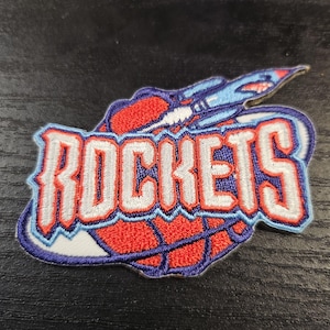 May include: Embroidered patch featuring the word "ROCKETS" in white, outlined in red and blue. The design includes a basketball and a rocket ship, with a blue and red color scheme. The patch has a white border.