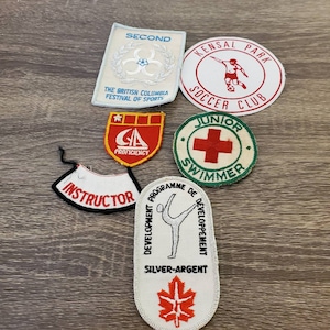 Peut inclure: Six patchs brodés. Les patchs sont blancs, rouges, bleus, verts et jaunes. Les patchs présentent divers motifs, notamment un joueur de football, un nageur, un gymnaste et une feuille d'érable. Les patchs portent le texte suivant : "Second", "The British Columbia Festival of Sports", "Kensal Park Soccer Club", "Junior Swimmer", "Instructor", "Development Programme de Developpement", et "Silver-Argent".