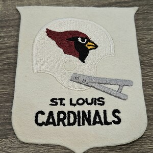 May include: White felt patch with a red cardinal wearing a white helmet with a silver face mask. The text "ST. LOUIS CARDINALS" is embroidered in black below the helmet.