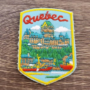 Puede incluir: Un parche bordado azul y amarillo con la palabra "Quebec" en letras rojas. El parche presenta una representación de un edificio con un techo verde y un cielo azul con nubes blancas. También hay pequeñas canoas rojas en el agua.