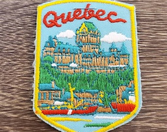May include: A blue and yellow embroidered patch with the word "Quebec" in red lettering. The patch features a depiction of a building with a green roof and a blue sky with white clouds. There are also small red canoes in the water.