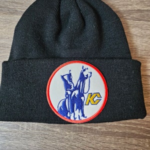 May include: Black knit beanie with a circular patch featuring a blue silhouette of a rider on a horse with the letters "KC" in yellow inside a red circle.