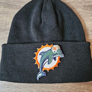 Peut inclure: Bonnet noir en tricot avec un grand patch brodé représentant un dauphin dans un soleil orange. Le dauphin est vert et bleu avec un contour blanc. Le soleil a des rayons orange et un centre blanc avec un "M" vert au centre.
