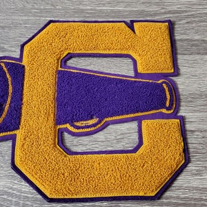 Op de afbeelding: Een gouden viltletter C met een paarse viltmegaphone erin. De letter is een patch of applicatie.