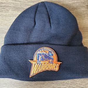 Puede incluir: Gorro negro con un parche del logotipo de los Golden State Warriors. El logotipo es un jugador de baloncesto en silueta azul y naranja con la palabra "Warriors" en naranja.