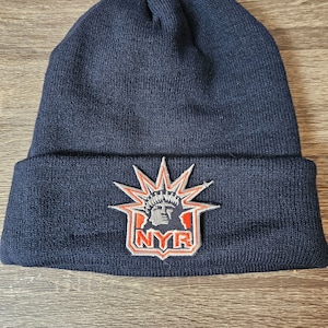 May include: A navy blue knitted beanie with a patch featuring the Statue of Liberty and the letters "NYR" in orange.
