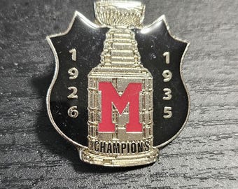 May include: A silver and black enamel pin featuring a stylised hockey trophy with the letter "M" and the years "1926" and "1935" on either side. The trophy has the word "CHAMPIONS" inscribed on the base.