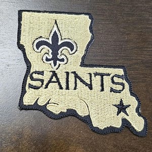 Op de afbeelding: Een beige geborduurde patch in de vorm van de staat Louisiana met een zwarte fleur-de-lis en het woord "SAINTS" in zwarte letters. Een zwarte ster bevindt zich in de rechterbenedenhoek van de patch.
