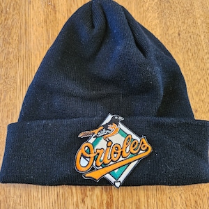 Może przedstawiać: Czarna czapka z dzianiny z haftowanym logo Baltimore Orioles przedstawiającym pomarańczowego ptaka i nazwę drużyny w pomarańczowych i białych literach na zielonym rombie.
