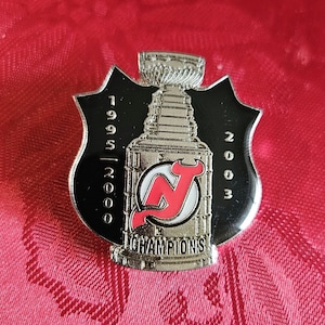 May include: A silver and black pin with a hockey team logo and the text "1995-2000 Champions 2003".