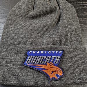 Może przedstawiać: Szara dzianinowa czapka z naszywką z logo Charlotte Bobcats. Naszywka ma niebiesko-pomarańczową kolorystykę z nazwą drużyny i wzorem głowy rysia. Czapka wykonana jest z miękkiego, dzianinowego materiału.