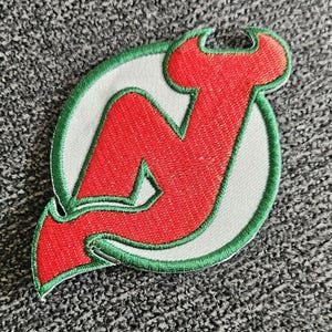 Könnte beinhalten: Ein gestickter Aufnäher mit dem Logo der New Jersey Devils. Das Logo ist ein stilisiertes "N" mit Flammen oben, in Rot und grün umrandet.