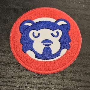 Peut inclure: Écusson brodé circulaire avec un visage d'ours bleu sur fond rouge. L'ours a des détails blancs et un design simple, de type dessin animé. Ce patch pourrait être pour des vêtements ou des accessoires.